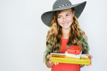 Beautiful teen girl in hat and red dress posing on grey background. Holiday shopping concept. Girl with long curly hair smiling and holding a gift boxes in her hands.