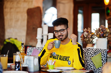 Asian man in eyewear and yellow sweatshirt sitting in cafe and drink coffee.