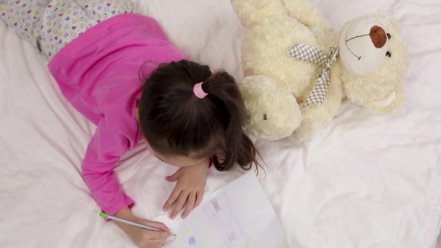 cute little girl drawing pictures while lying on bed. Kid painting at home. top view