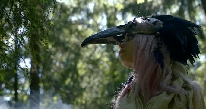 young female shaman is wearing bird skull mask on head, standing in daytime in cloud of smoke