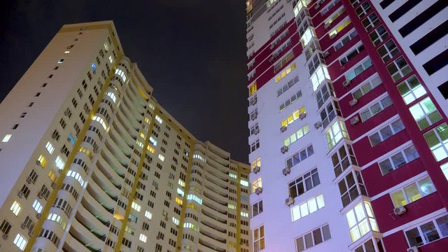 Nighttime time lapse of illuminated building windows at night with people living in flats with balcony. Electricity consumption concept. View from below. City street in evening