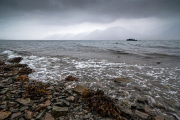 Sea bay in the rain. Severe seascape with coast and mountains. Boat in the sea. Surf and seaweed. Autumn weather. Arctic nature. Egvekinot Bay, Kresta Gulf, Bering Sea, Chukotka, Far East of Russia.