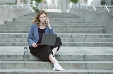 Business woman working with phone device and laptop computer outdoors near the office on the stairs. Freelance worker is busy talking via phone holding the laptop sitting on the stairs.