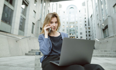 Portrait of a beautiful businesswoman sitting on the stairs talking via phone with the employee holding her laptop. Young businessperson looking at the camera holding her phone, computer and water.