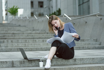 Concentrated young businesswoman reading the documents sitting on the stairs near the business center. Laptop and coffee near the busy diligent employee attentively reading the papers.