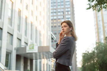 Fototapeta premium Thoughtful businesswoman with wireless headphones posing in the street near office buildings and skyscrapers. Young businessperson in earphones in front of business center. Pensive, concentrated look.