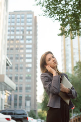 Young successful businesswoman in smart casual clothes in front of office buildings and skyscrapers. Woman holding modern laptop computer, listening to music in wireless headphones on street outdoors.