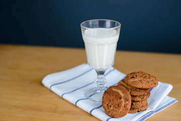 round brown wholemeal oatmeal cookies stack with raisins and white milk in glass cup on striped textile rag on wooden table, close up side view of vertical still life stock photo image