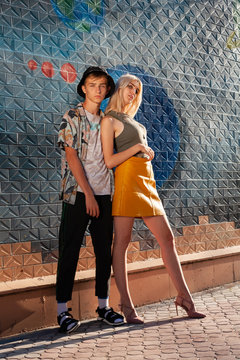 Young Guy With A Girl Stylishly Dressed In Fashion. Against The Background Of The Wall. The Guy In The Shirt And T-shirt With A Floral Print. Sandals With Socks.