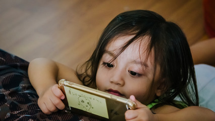 Bangi, Malaysia - July 1, 2019: Adorable Asian toddler girl laying on the floor and playing with mobile phone at home, indoors. Child with tablet computer.