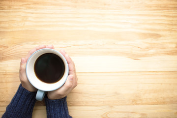 A woman wearing a blue jersey holding a cup of coffee with her hands. Place it on the wood table on the left hand side of the picture. Top view.