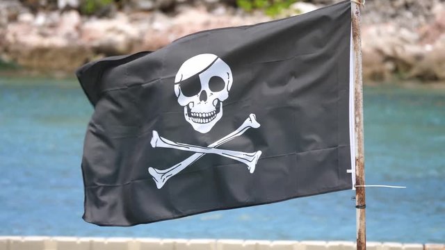 A black and white pirate skull and bones flag mounted to a weathered and rusted steel post flaps in the wind with blue water and rocky shoreline in the background.