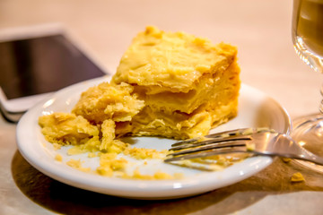 A half-eaten slice of Napoleon cake on a saucer, a mobile phone and a cup of cappuccino stand on a light wooden table.
