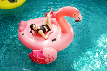 Young and sexy woman, having fun and laughing on an inflatable giant pink flamingo in the pool Float air mattress in a bikini.