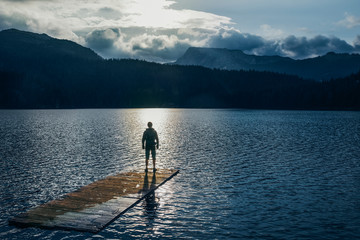 Naklejka premium Silhouette man standing on the wooden raft on the blue lake. Cloudy mountain in the back with the evening light in nature landscape. Reflexion on the raft. Black Lake, Montenegro, Durmitor