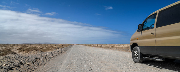 Fototapeta na wymiar Landscape of a typical white dirt road near El Coralejo Fuerteventura Canary Island Spain. 4x4 van along an offroad path, blue sky with clouds in background. Travel adventure and freedom concept.
