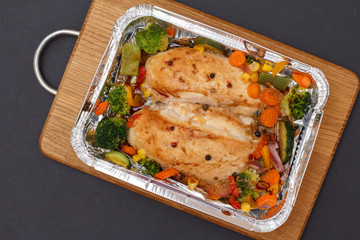 Baked chicken breasts or fillet in metal container with vegetables on cutting board.