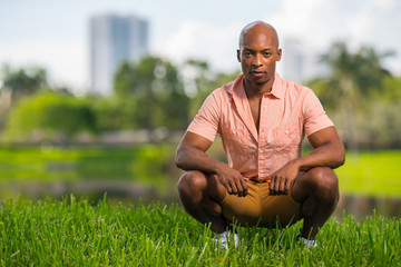 Naklejka na ściany i meble Photo of a handsome young African American man squatting on grass in the park. Deadpan expression staring deep into the camera