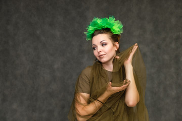 Portrait in lower chest on a gray background of a pretty young brunette woman with a green floral wreath in her hair. Standing in different poses, talking, showing hands, demonstrating emotions.