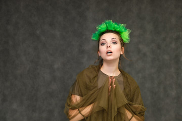 Portrait in lower chest on a gray background of a pretty young brunette woman with a green floral wreath in her hair. Standing in different poses, talking, showing hands, demonstrating emotions.