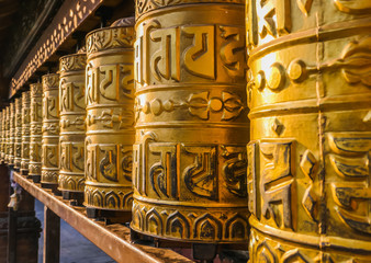 A prayer wheels on a spindle made from metal and wood. Mantra Om Mani Padme Hum is written in Newari language of Nepal on the outside of the wheels. Golden Temple (Kwa Bahal) buddhist temple in Patan - obrazy, fototapety, plakaty