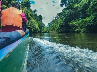 A boat tour in Amazonas. Water splashes on the side, as the boat goes forward. On both sides of the river thick mangrove jungle. One man sitting on the boat, wearing orange life jacket.