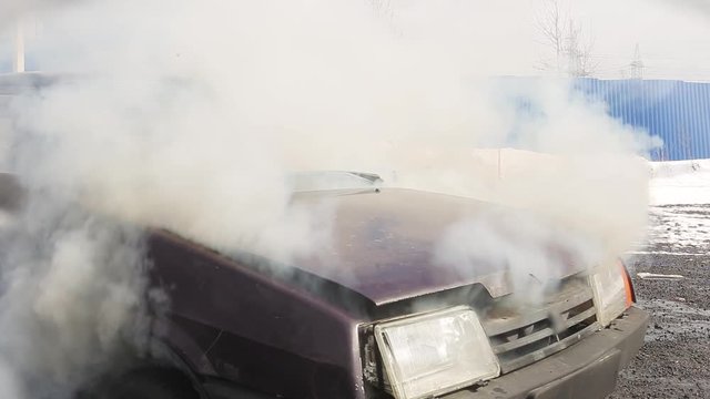 Abandoned Rusty Dirty Old Dark Dusty Violet Car Is Left On Deserted Grey Road At Some Suburb Area Where Car's Closed Hood Is Burning And Slightly Smoking On Cold Cloudy Winter Day.