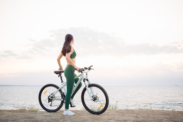 Fototapeta premium Beautiful young brunette woman resting after bicycle ride at sunrise. Model listening to music with wireless earbuds in the morning.