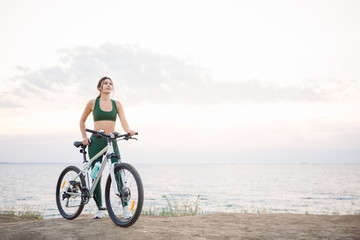 Fototapeta premium Beautiful young brunette woman resting after bicycle ride at sunrise. Model listening to music with wireless earbuds in the morning.