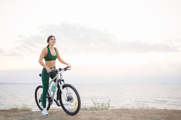 Beautiful young brunette woman resting after bicycle ride at sunrise. Model listening to music with wireless earbuds in the morning.