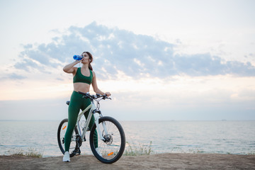 Fototapeta premium Young brunette woman with wireless earbuds and smart watches resting after bicycle ride at sunrise drinking water.