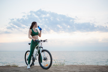 Young brunette woman with wireless earbuds and smart watches resting after bicycle ride at sunrise drinking water.