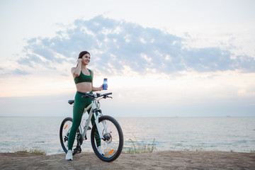 Fototapeta na wymiar Young brunette woman with wireless earbuds and smart watches resting after bicycle ride at sunrise drinking water.