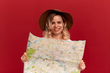 Blond girl with curly hair in a white t-shirt and a sundown hat looks at the map looking for itinerary while traveling.