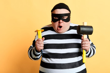 angry plump criminal in striped clothes preparing for crime, isolated yellow background. studio shot