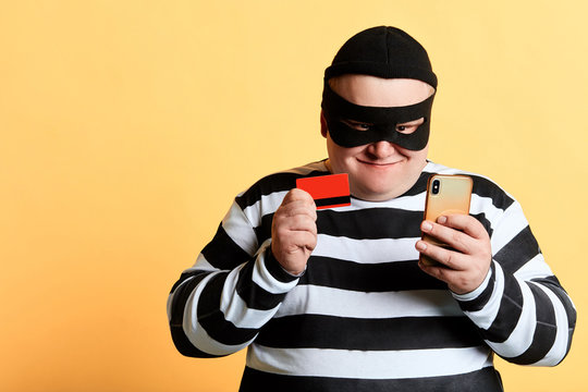happy cheerful plump hacker with a bank card and mobile phone Isolated on the yellow Background. crime, illegal business copy space, isolated yellow background