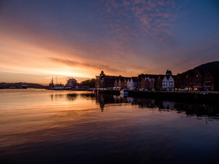 Beautiful front line of houses build in the port in Bergen, Norway. Soft reflections of houses and boats in the sea. Behind the buildings there is a small hill. Golden hour, sunset in the port.