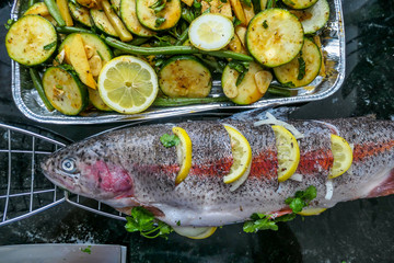 A salmon stuffed with vegetables and lemon placed in a fish grilling basket. On the side a plate with sliced lemons, tomatoes and green beans ready to be grilled. Healthy lifestyle meal.