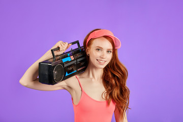 red-haired young woman with freckles looks in camera with smile, holds stereo and cassette tape...
