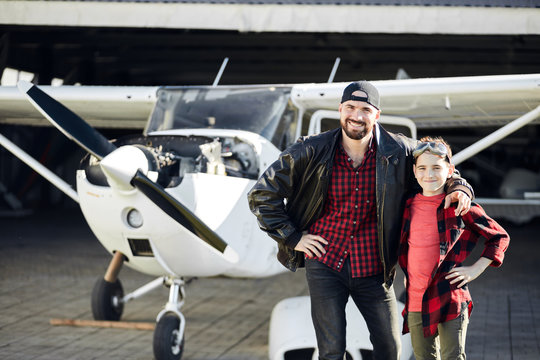 Boy in aviator glasses stands wih his dad, proud of his father pilot, wants to be like him when he is grown up, both looking in camera with smile, propeller plane stands on background, outdoor shot.