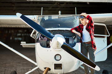 adorable boy in aviator glasses stands beside small white propeller airplane, scratches his head in...