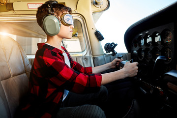 side view shot of teenager boy in pilot headset and aviator glasses, sitting in airplane cabin,...