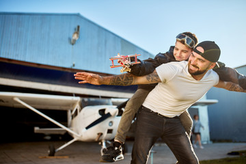 view of smiling boy in large leather pilot jacket with small model plane, riding on father's back, playing outside hangar after the flight on their light private propeller airplane. © alfa27