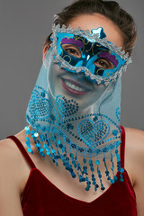 Half-turn shot of smiling woman with dark hair, wearing wine red crop top. The girl is tilting her head, wearing blue glossy masquerade mask, adorned with long veil with paillettes and ladder braid.