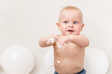 Baby birthday party. Child eating birthday cake. The boy on a light background with white ballons and copy space smash the cake