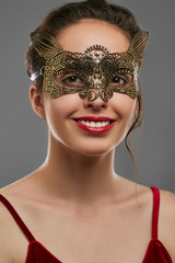 Half-turn shot of smiling lady with dark hair, wearing wine red crop top. The woman is looking at the camera, wearing golden carnival mask with perforation and jutting edges, tied with satin ribbon.