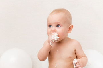 Baby birthday party. Infant eating birthday cake. The boy on a light background with white ballons and copy space smash the cake