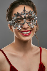 Half-turn shot of smiling lady, wearing wine red crop top. The young woman is tilting head, wearing asymmetric silver carnival mask with perforation and cross on forehead, tied with satin ribbon.