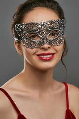 Portrait of smiling woman with tied back dark hair, wearing wine red crop top. The young girl is tilting her head, wearing silver carnival mask with perforation. Vintage women's carnival accessory.