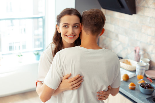 Appealing dark-eyed wife hugging husband in the kitchen